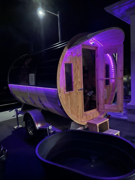 The barrel sauna at night with its door open and purple lights on, cold plunge tub in front
